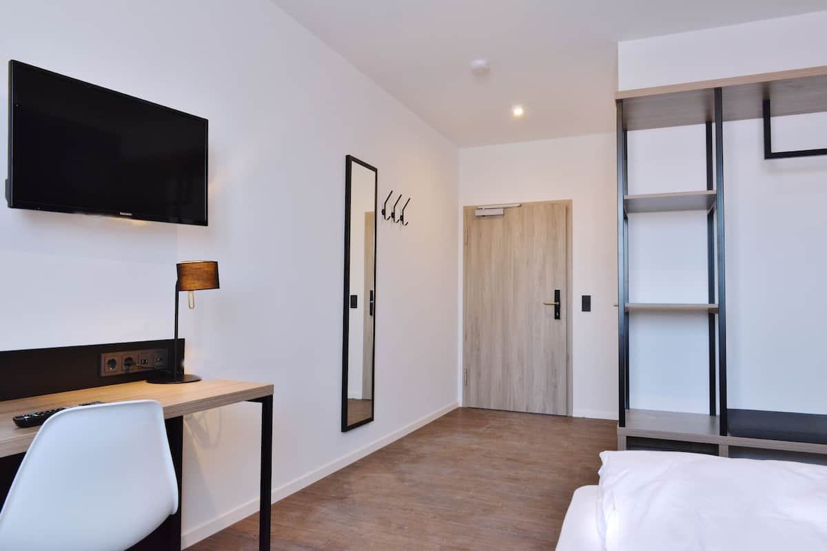 A modern hotel room with a flat-screen TV, a desk with a lamp, a chair, a bed, a wardrobe, and a door.