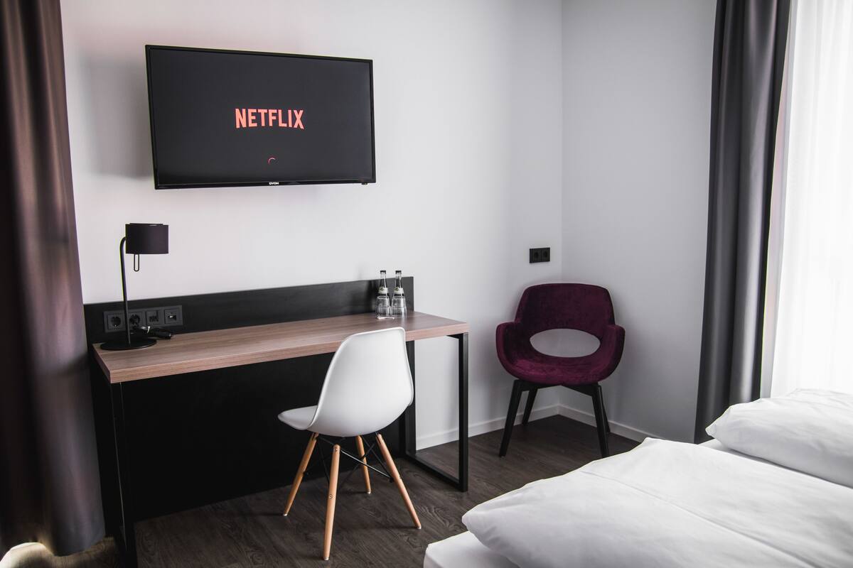 A hotel room with a bed, a desk with a chair, a TV displaying 'NETFLIX', and a window with curtains.