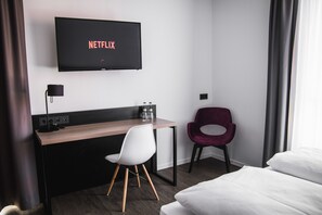 A hotel room with a bed, a desk with a chair, a TV displaying 'NETFLIX', and a window with curtains. - Hayta Hotel Stuttgart Airport Messe (Filderstadt)
