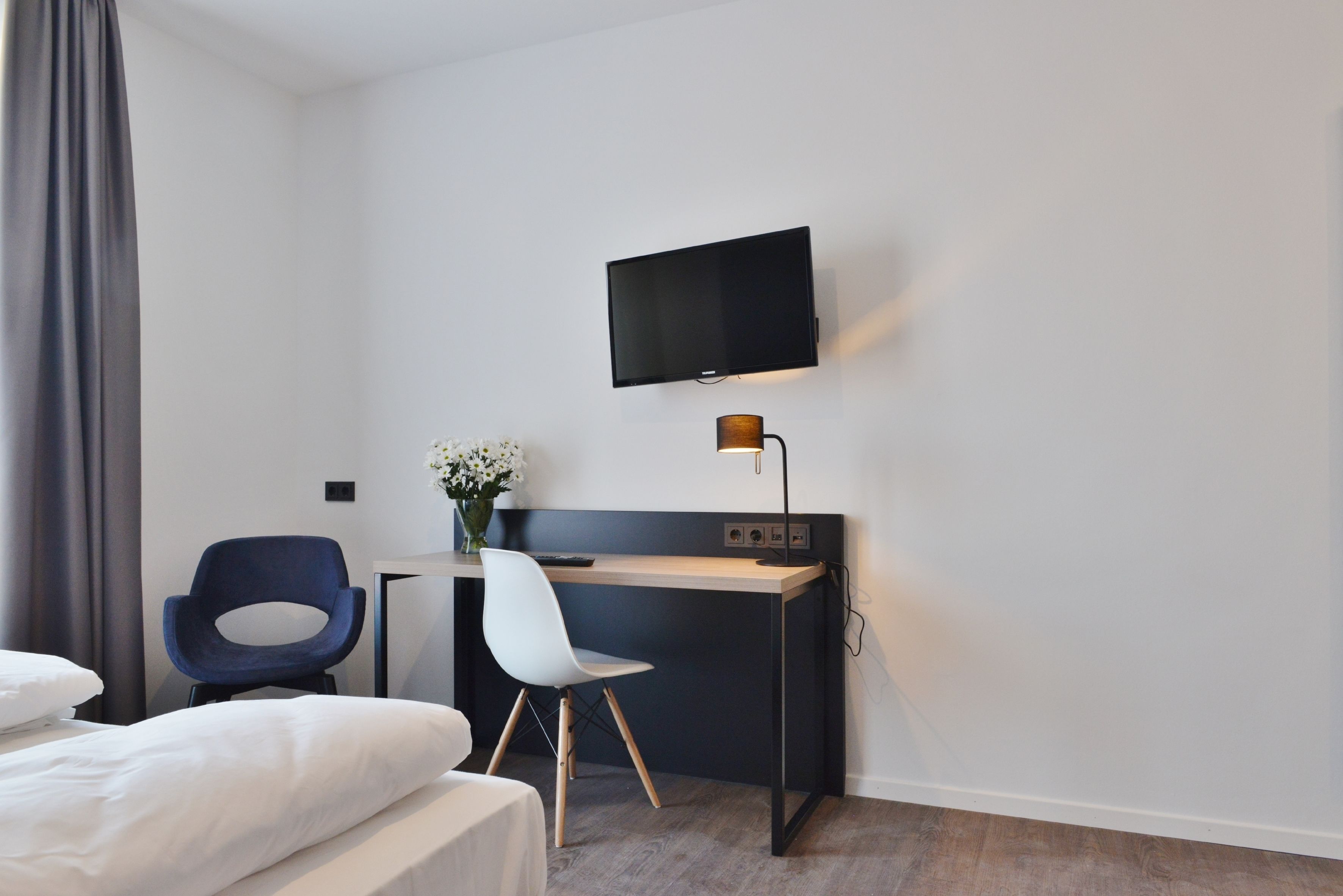 A hotel room with a bed, a desk with a chair, a flat-screen TV mounted on the wall, and a desk lamp.