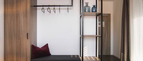 A bedroom with a bed, a wooden wardrobe, a shelf with items, and a bench with a pillow.