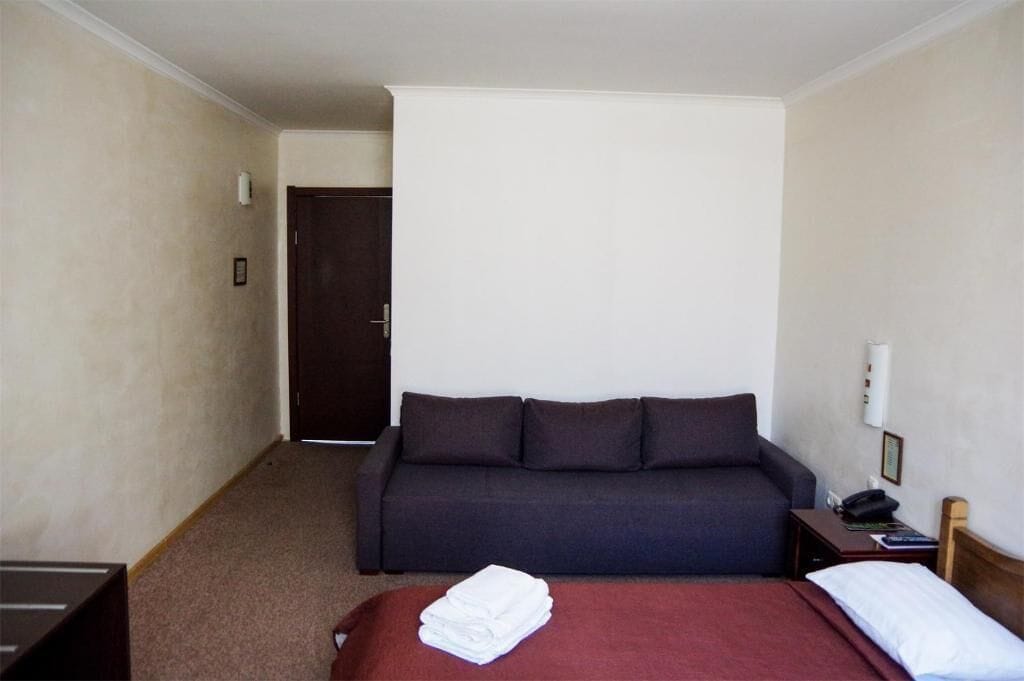 Habitación de hotel con cama, sofá de color oscuro, mesita de noche de madera con lámpara, y una puerta que da a otra habitación.