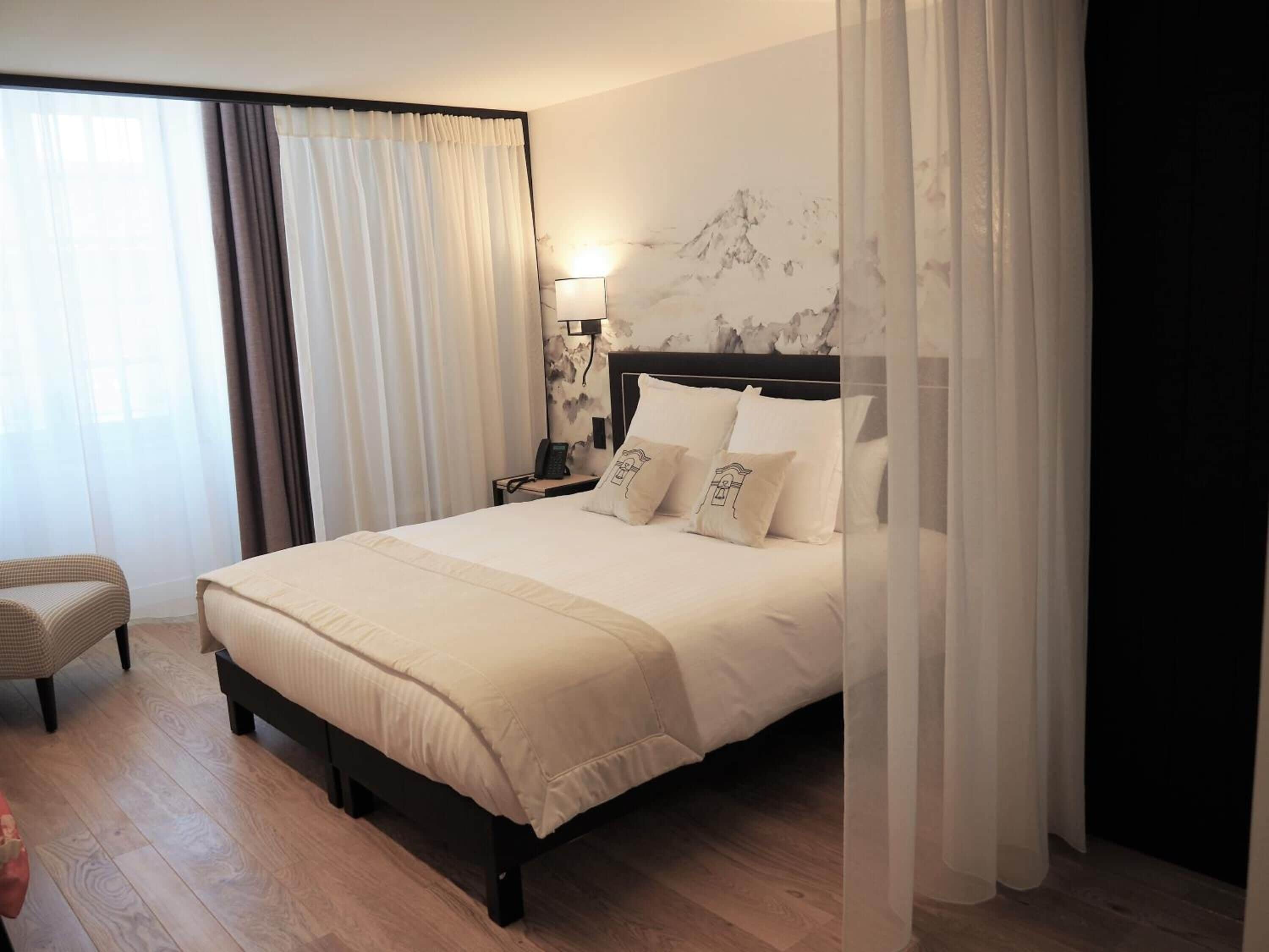 A hotel room with a large bed, a bedside table with a lamp, a chair, and a mural on the wall.