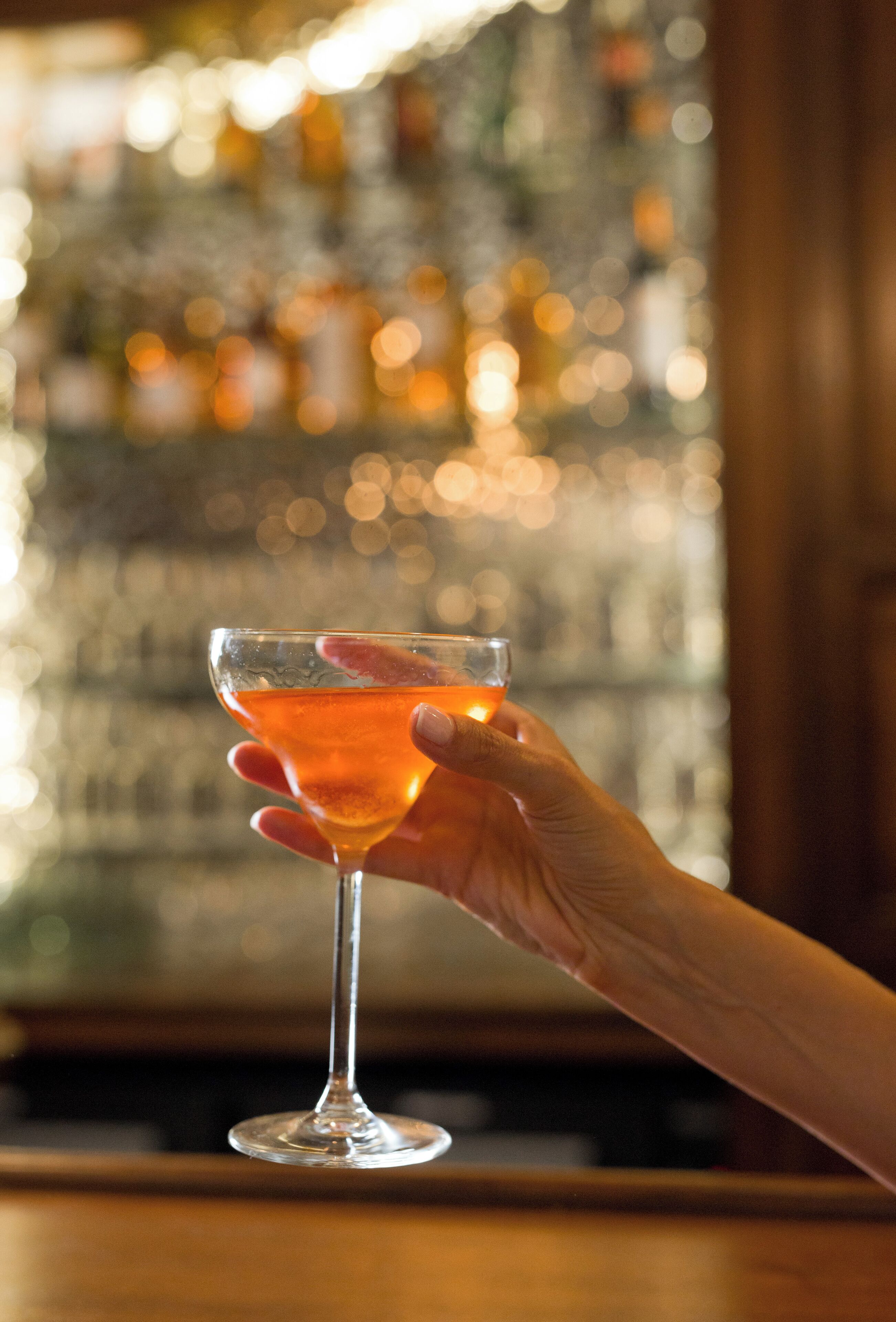 A hand holding a cocktail glass with a blurred background of a bar and bottles.