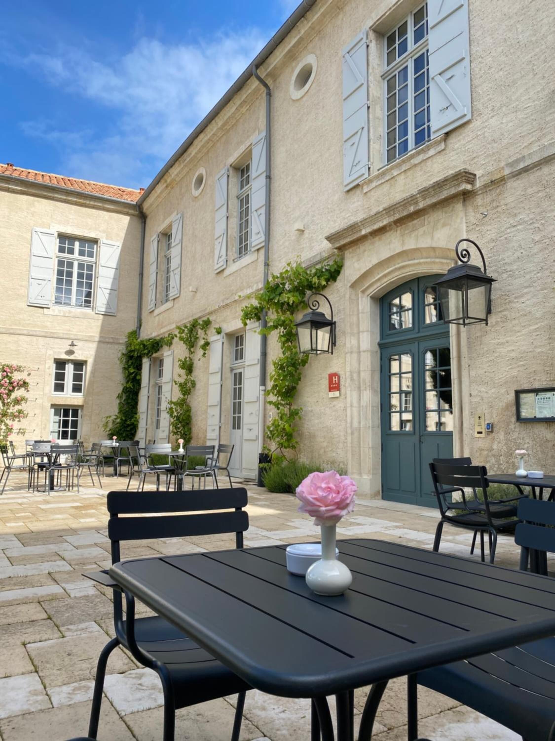 An outdoor café area with tables and chairs, a decorative flower vase, and a building with a green door and white shutters.