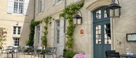 An outdoor café area with tables and chairs, a decorative flower vase, and a building with a green door and white shutters.
