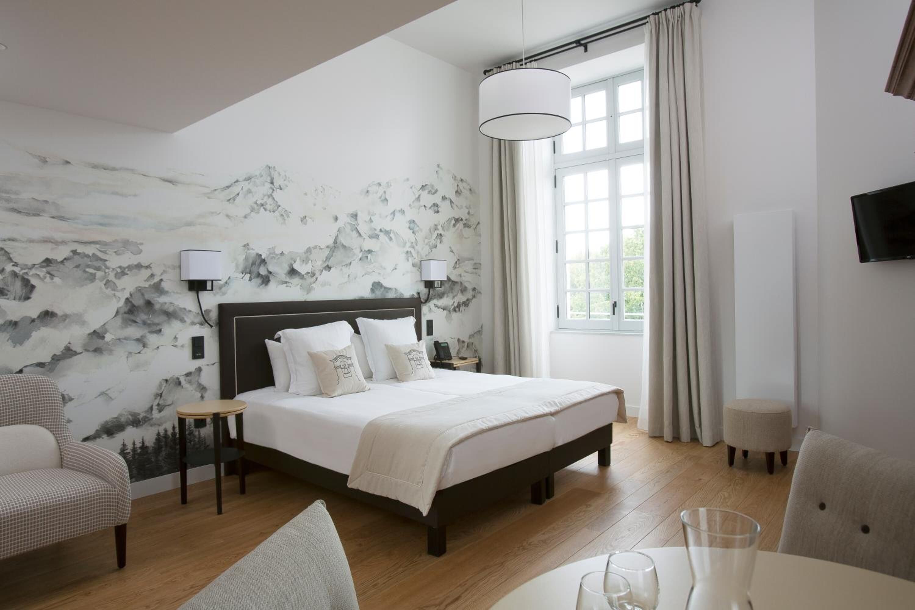 Une chambre d’hôtel comprenant un lit, deux tables de chevet, une chaise, une petite table, un téléviseur et une fenêtre avec des rideaux.