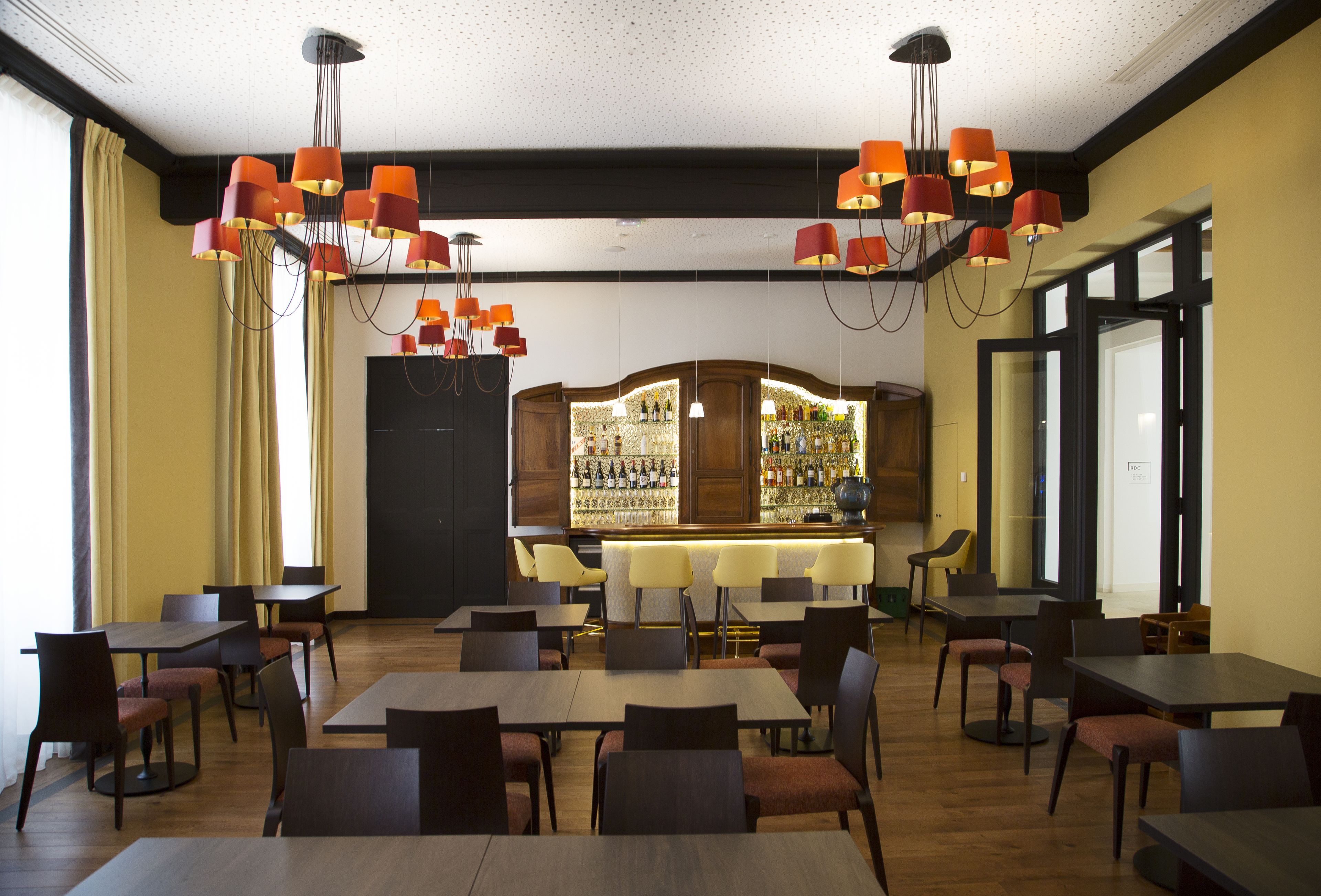 Un restaurante con varias mesas y sillas, una zona de bar con botellas, y un ventanal grande al lado izquierdo.