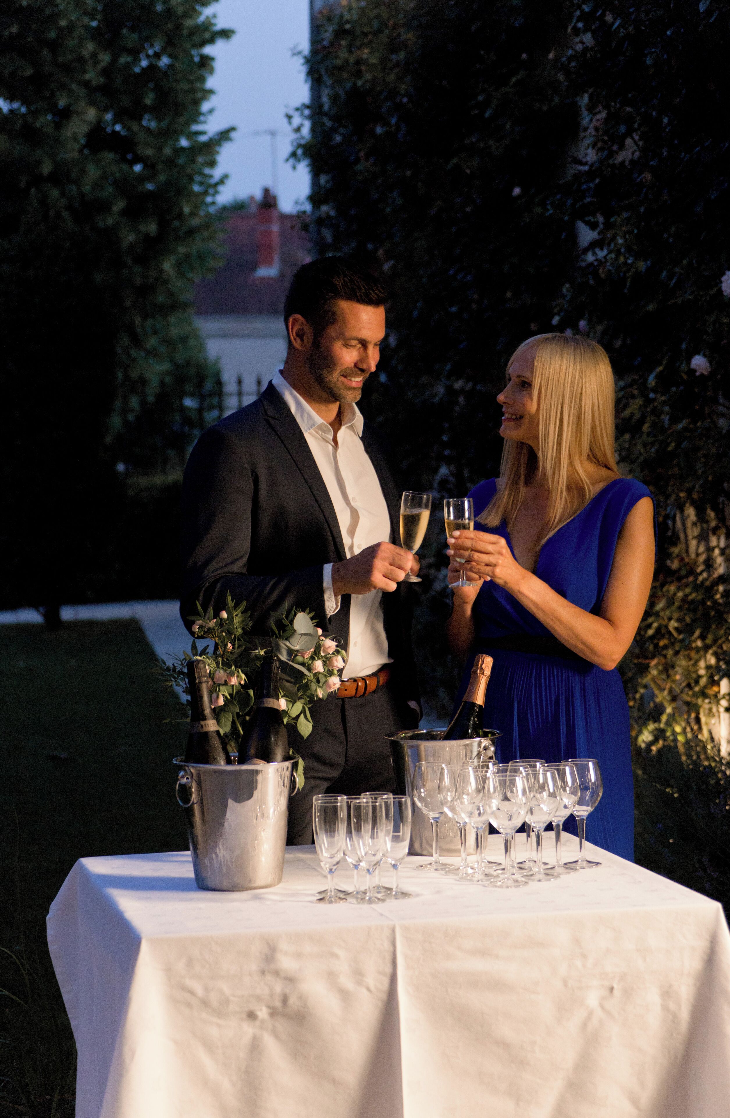 Una pareja brindando con copas de champán en una mesa al aire libre con mantel blanco, rodeada de árboles.