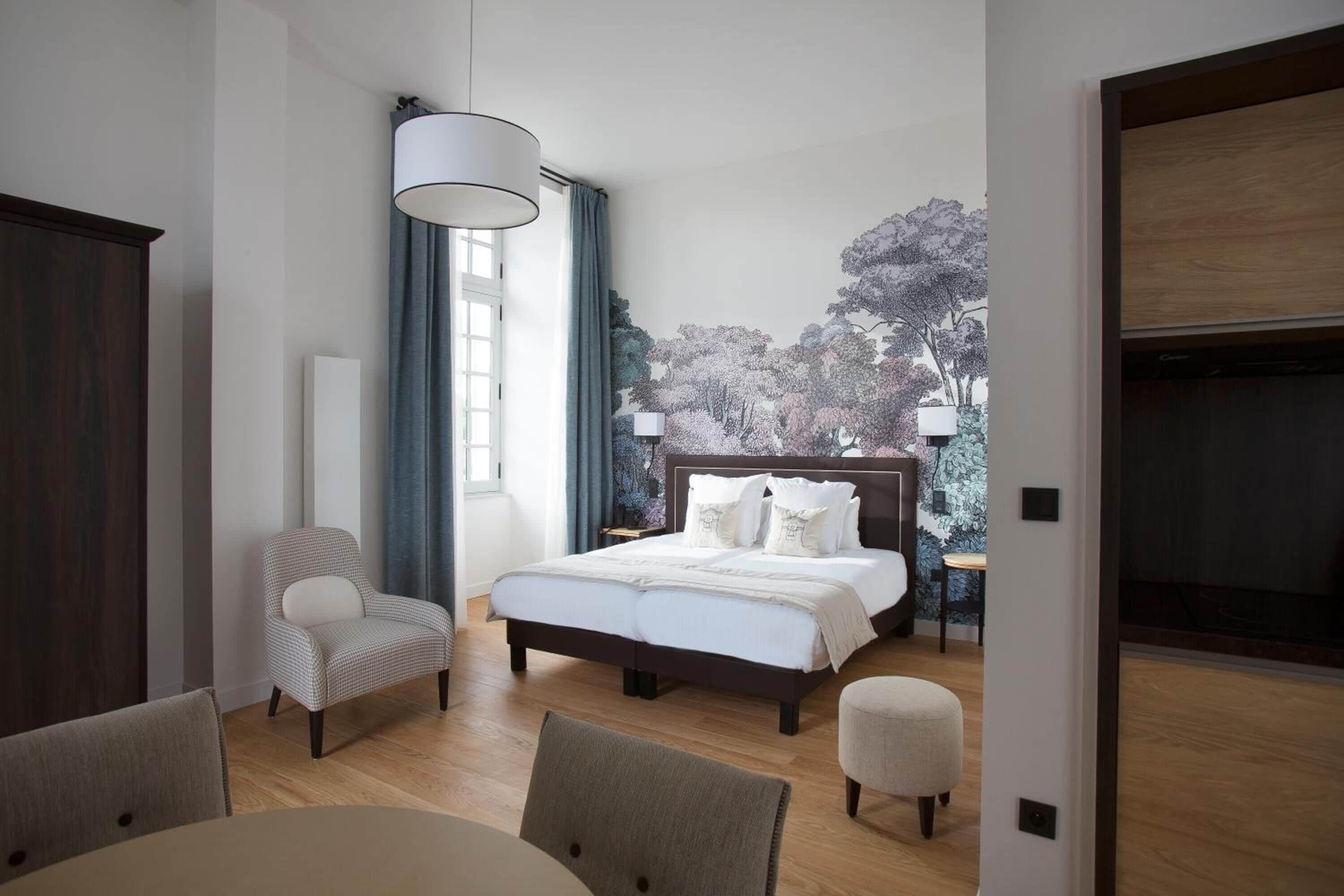 A modern bedroom with a large bed, a chair, a table, and a view of trees through the window.