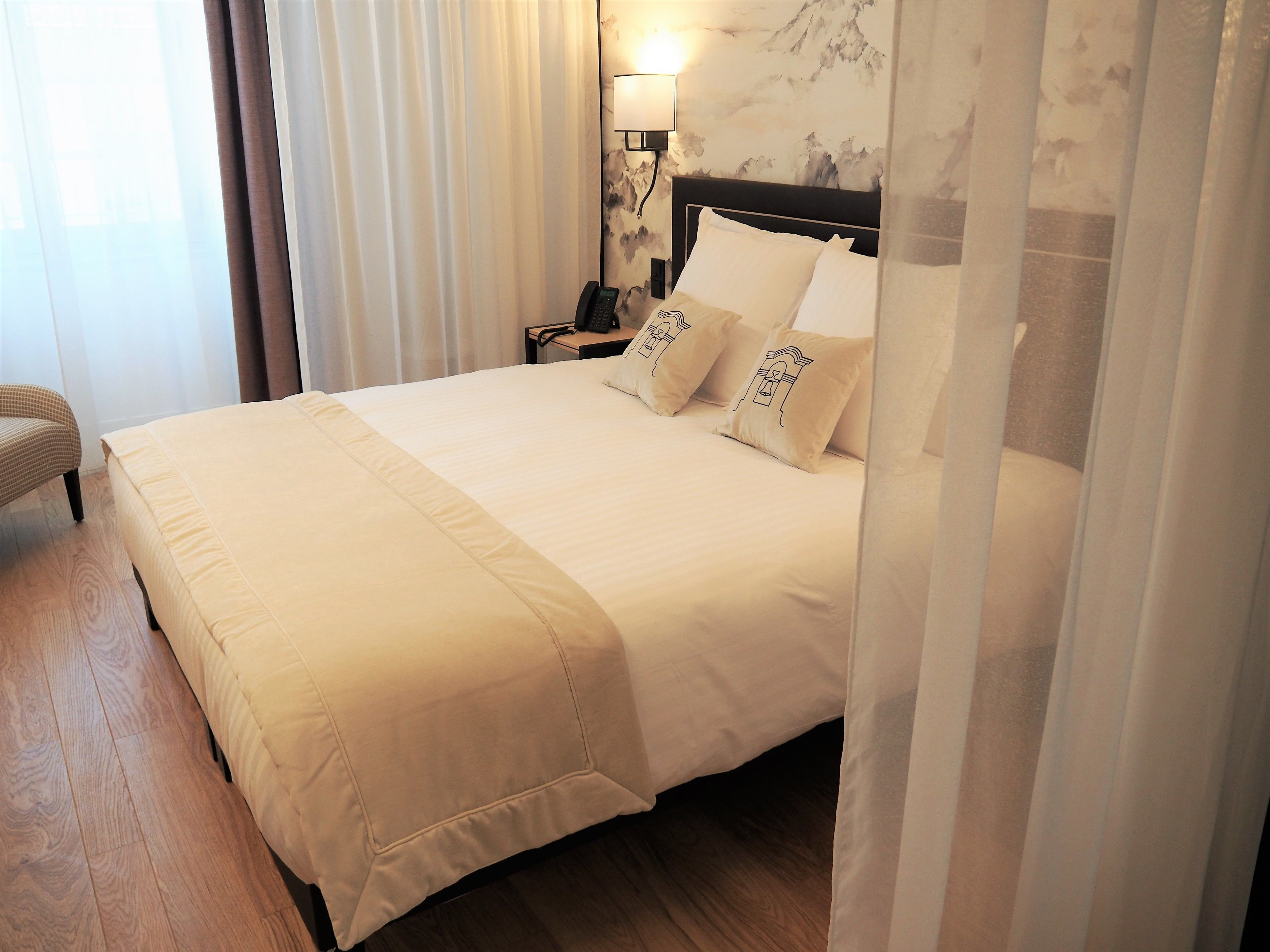A neatly made bed with a white duvet and pillows, a bedside table with a lamp, and a chair in a hotel room.