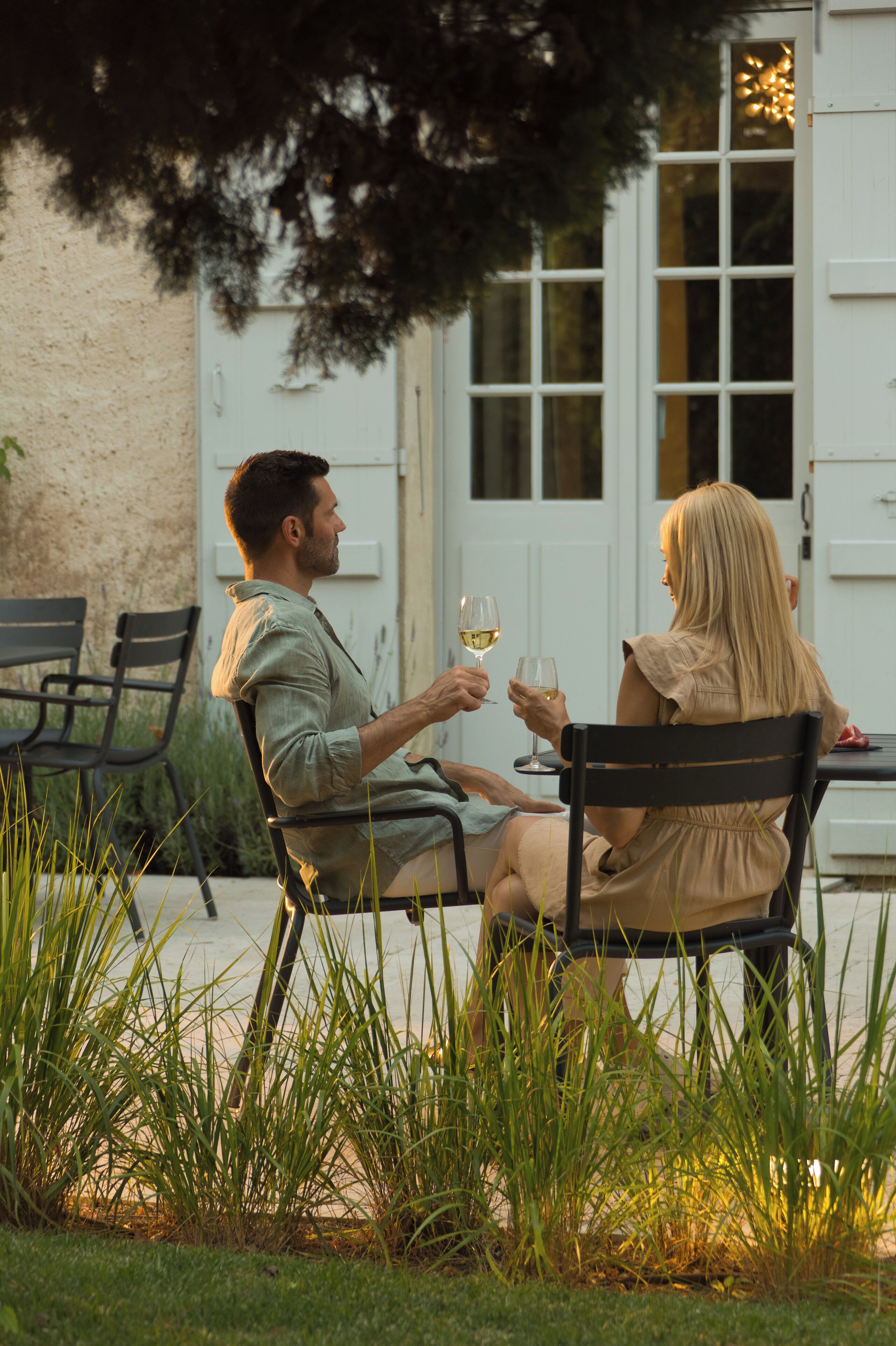 Two people seated at an outdoor table with chairs, holding wine glasses.