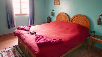 Une chambre à coucher avec un lit, une fenêtre avec des rideaux, un ventilateur fixé au mur et une tête de lit décorative.