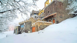 Un bâtiment historique en pierre, doté de nombreuses portes et fenêtres en bois, entouré d’arbres couverts de neige.