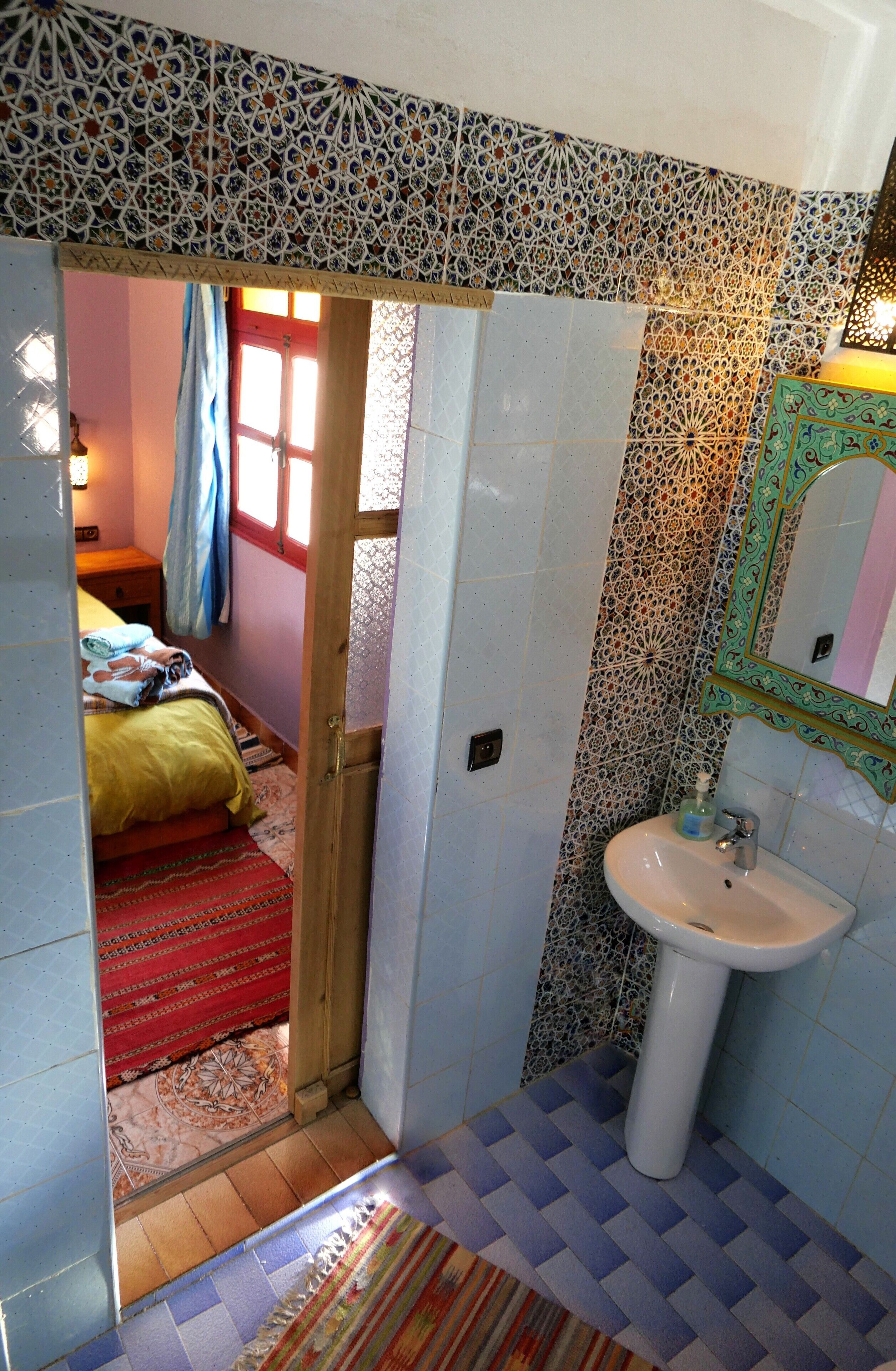Un baño con lavamanos, baldosas estampadas y un acceso a un dormitorio con cama y ventana.