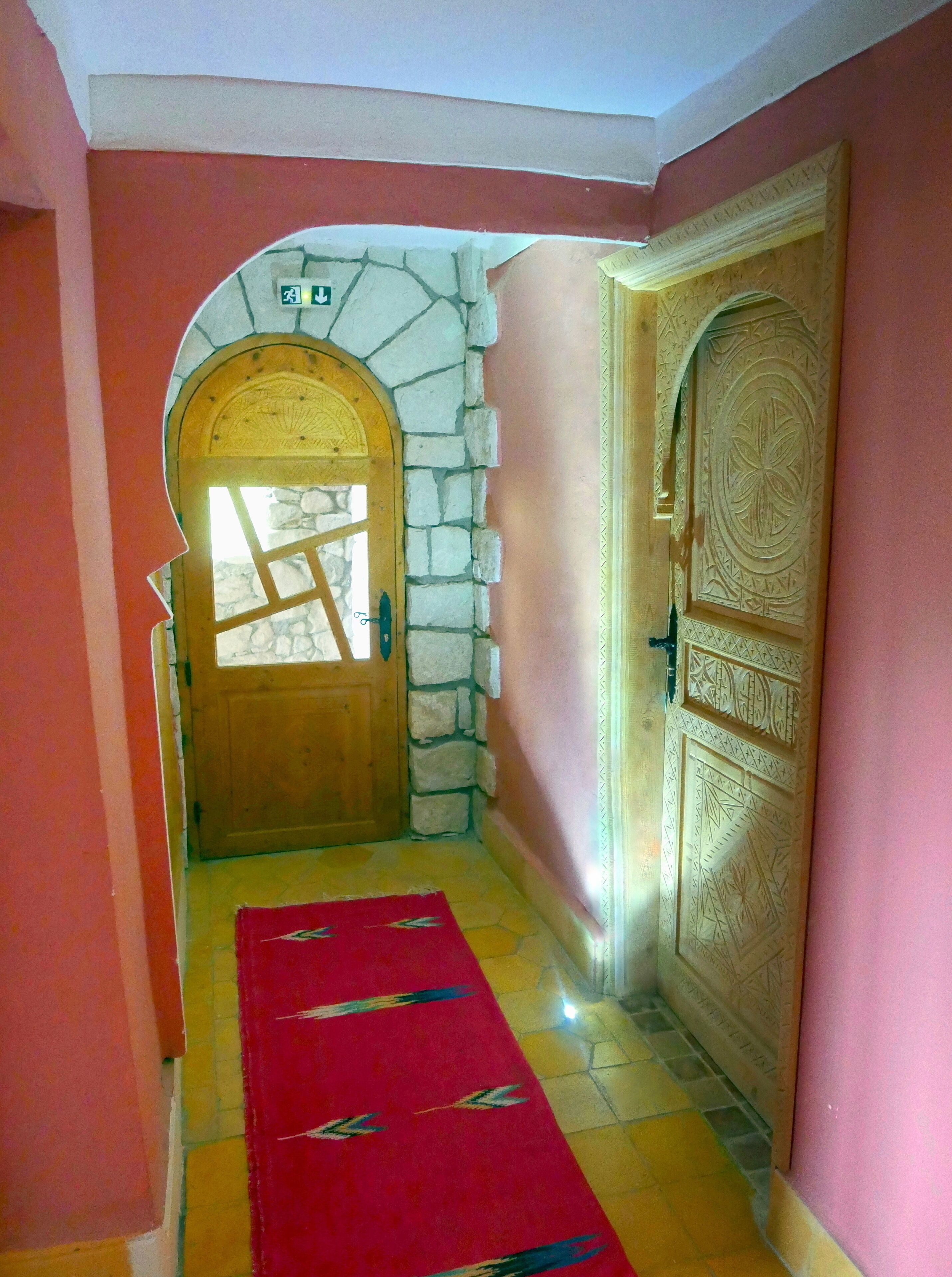 Un pasillo con alfombra roja, una puerta de madera y un arco de piedra, y una pared con un panel decorativo.