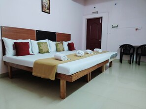 Habitación de hotel con una cama grande, cabecera de madera y un banco al pie de la cama. Hay dos sillas contra la pared y una puerta con pomo.