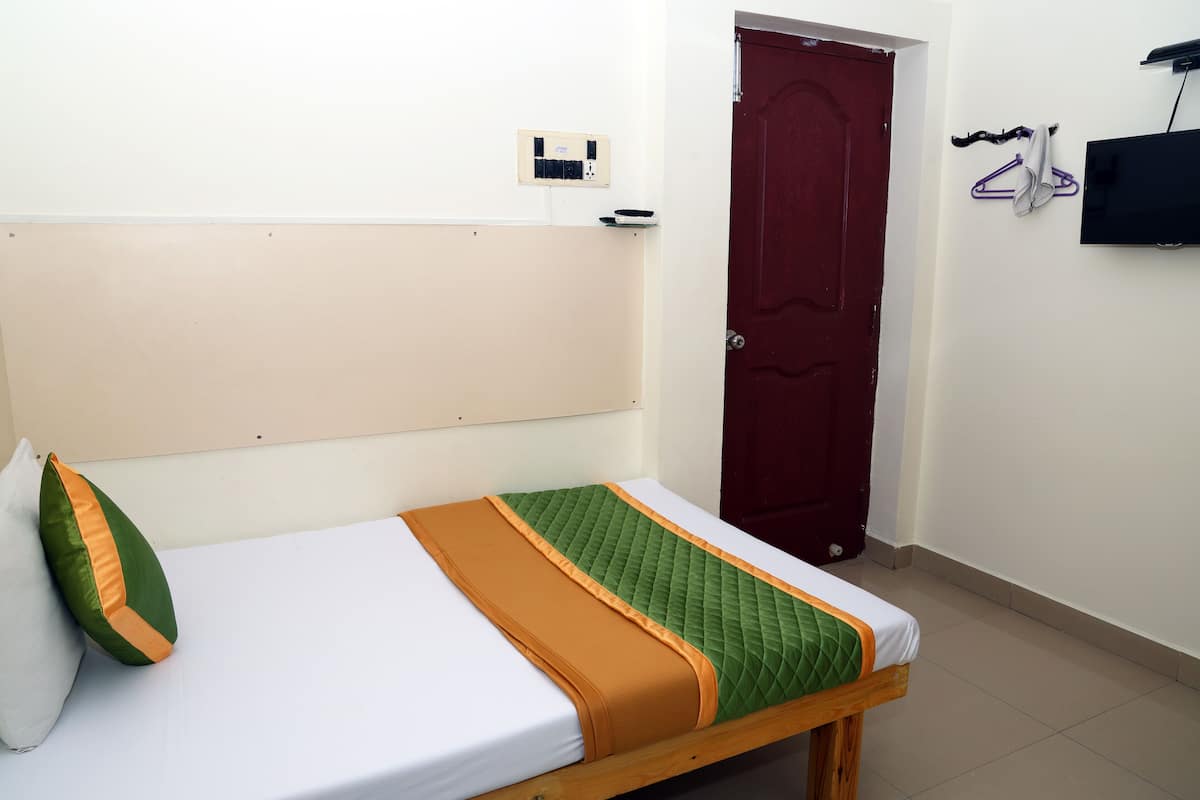 Un lit simple avec une couette verte, une couvre-lit orange et un oreiller blanc. On aperçoit une porte rouge au fond.
