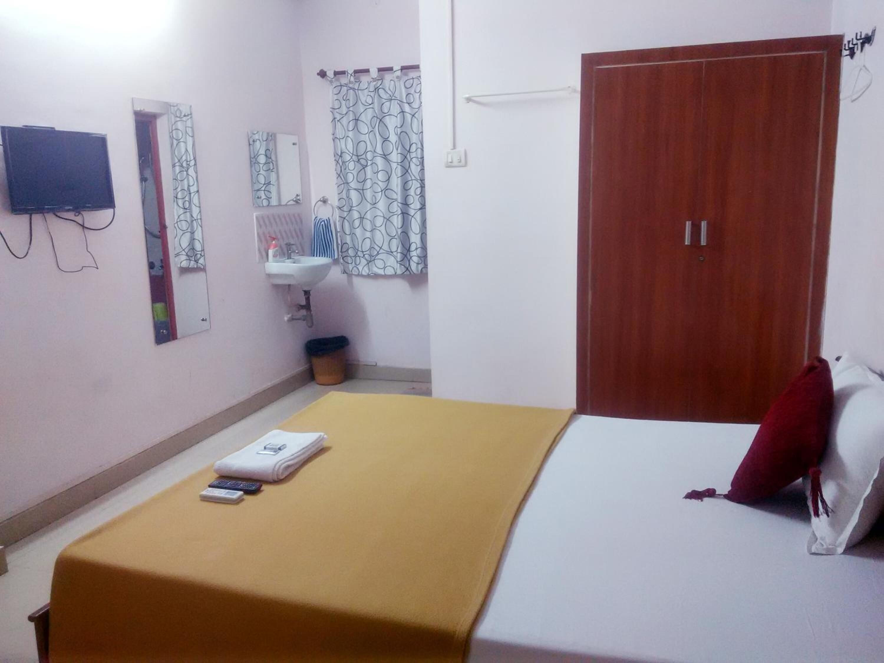 Un dormitorio con cama, una mesita de noche con un libro y un control remoto, un armario de madera, un baño con lavamanos y espejo, y un televisor montado en la pared.