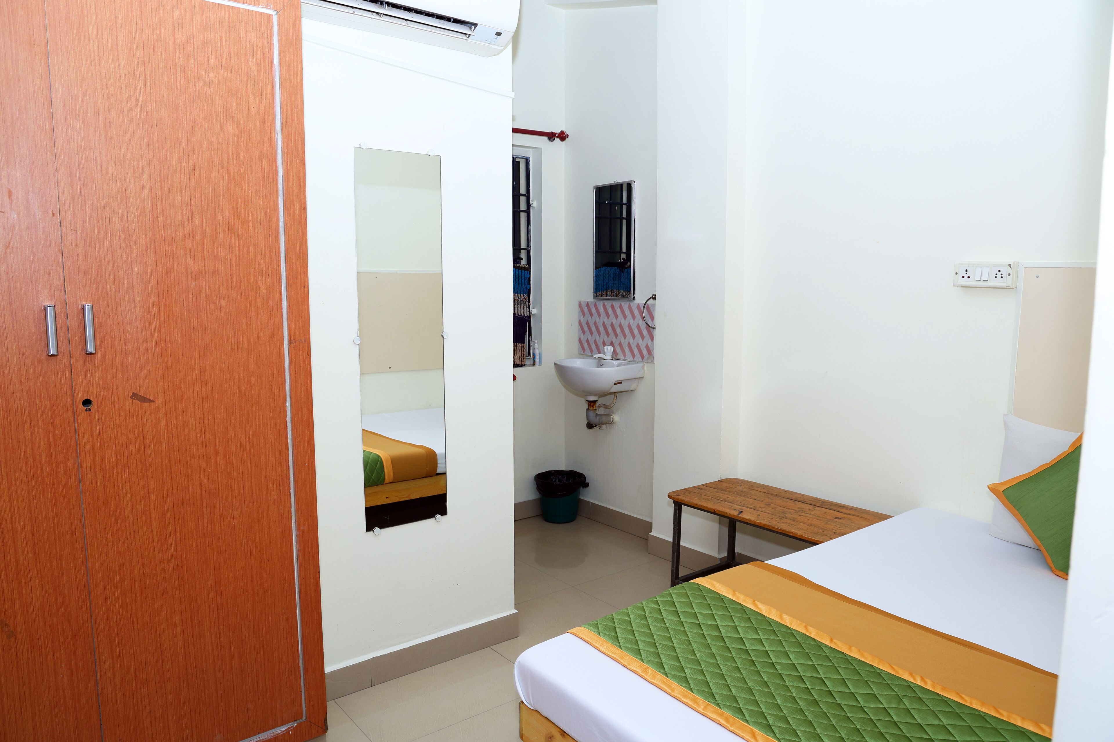 A small, clean room with a bed, a wooden wardrobe, a small table, a sink, and a mirror.