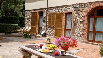 Un bâtiment en pierre avec des volets en bois, une terrasse où est dressée une table pour le petit-déjeuner, et un jardin parsemé de fleurs jaunes.