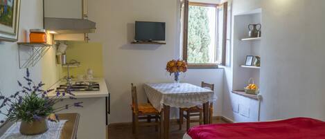 Ein kleiner, hell erleuchteter Raum mit einem Bett, einem Tisch mit Blumenvase, einem Fernseher an der Wand und einem Fenster mit Blick auf Bäume.