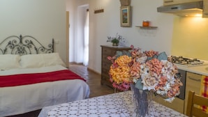 A bedroom with a bed, a nightstand, and a table with a vase of flowers. There is a kitchen area with a stove and cabinets. - Casa Vacanze Dolce Vita (Campiglia Marittima)