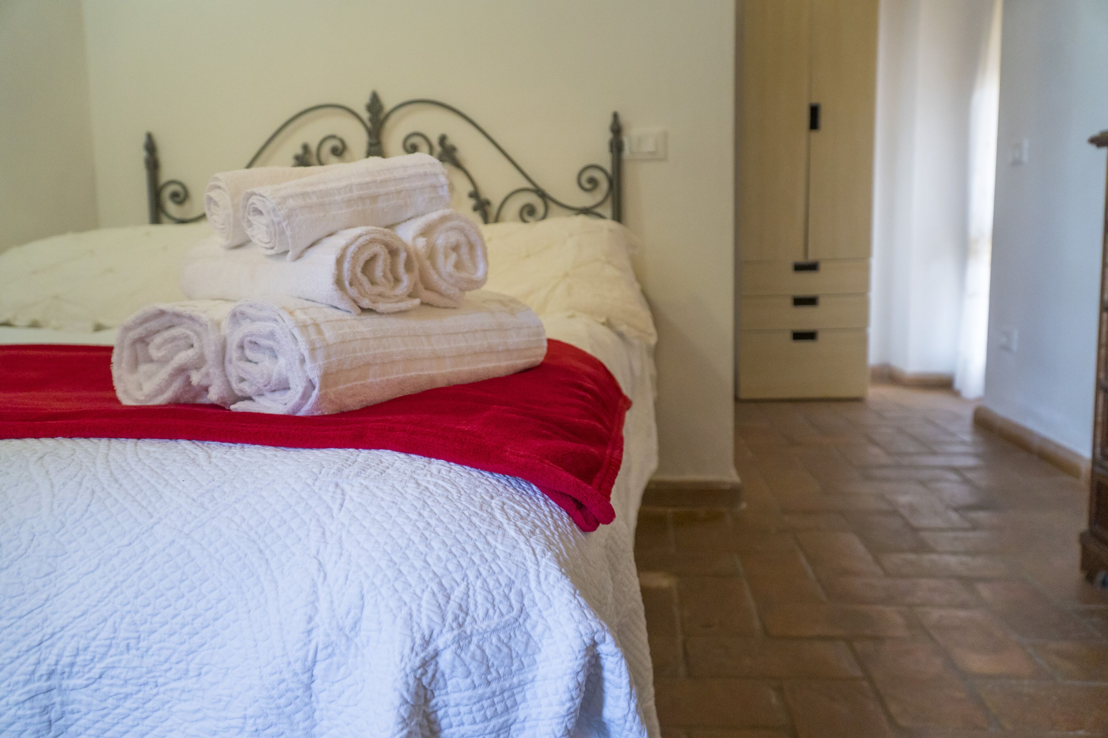 Een keurig opgemaakt bed met opgerolde handdoeken, een rode deken, een wit sprei en een metalen hoofdbord.
