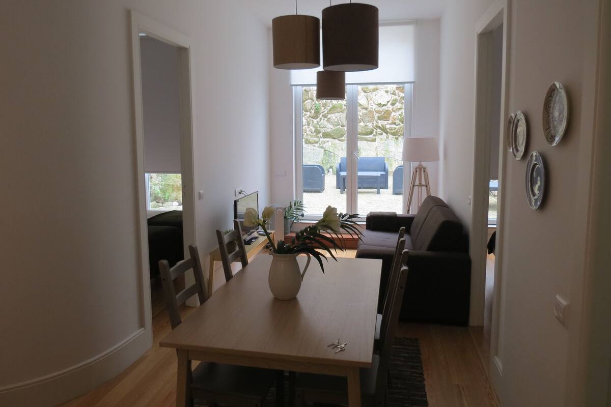 A dining area with a wooden table, chairs, a vase with flowers, and a view of the outdoors through large windows.