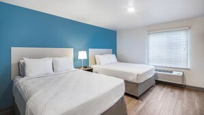 A hotel room with two beds, a blue accent wall, a window with blinds, and a built-in heater. - WoodSpring Suites Chicago Addison (Addison)