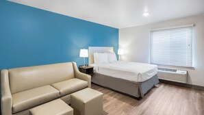 A hotel room with a bed, a sofa, a nightstand with lamps, and a window with blinds. - WoodSpring Suites Chicago Addison (Addison)