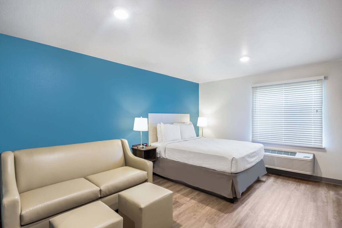 A hotel room with a bed, a sofa, a nightstand with lamps, and a window with blinds.