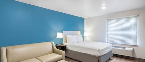 Quarto de hotel com cama, sofá, mesa de cabeceira com luminárias e janela com persianas.