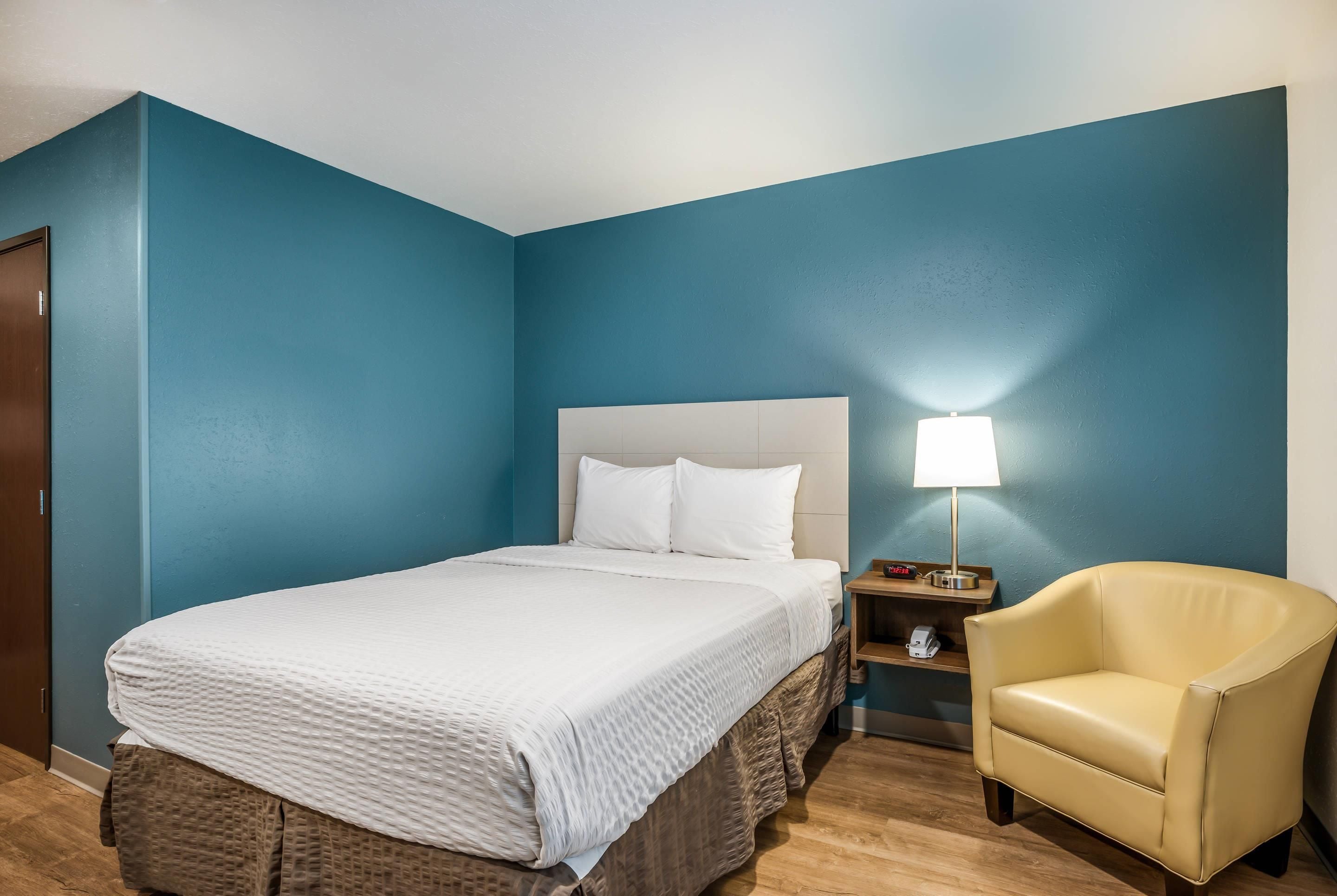 Quarto de hotel com cama, cadeira, mesa de cabeceira e abajur.