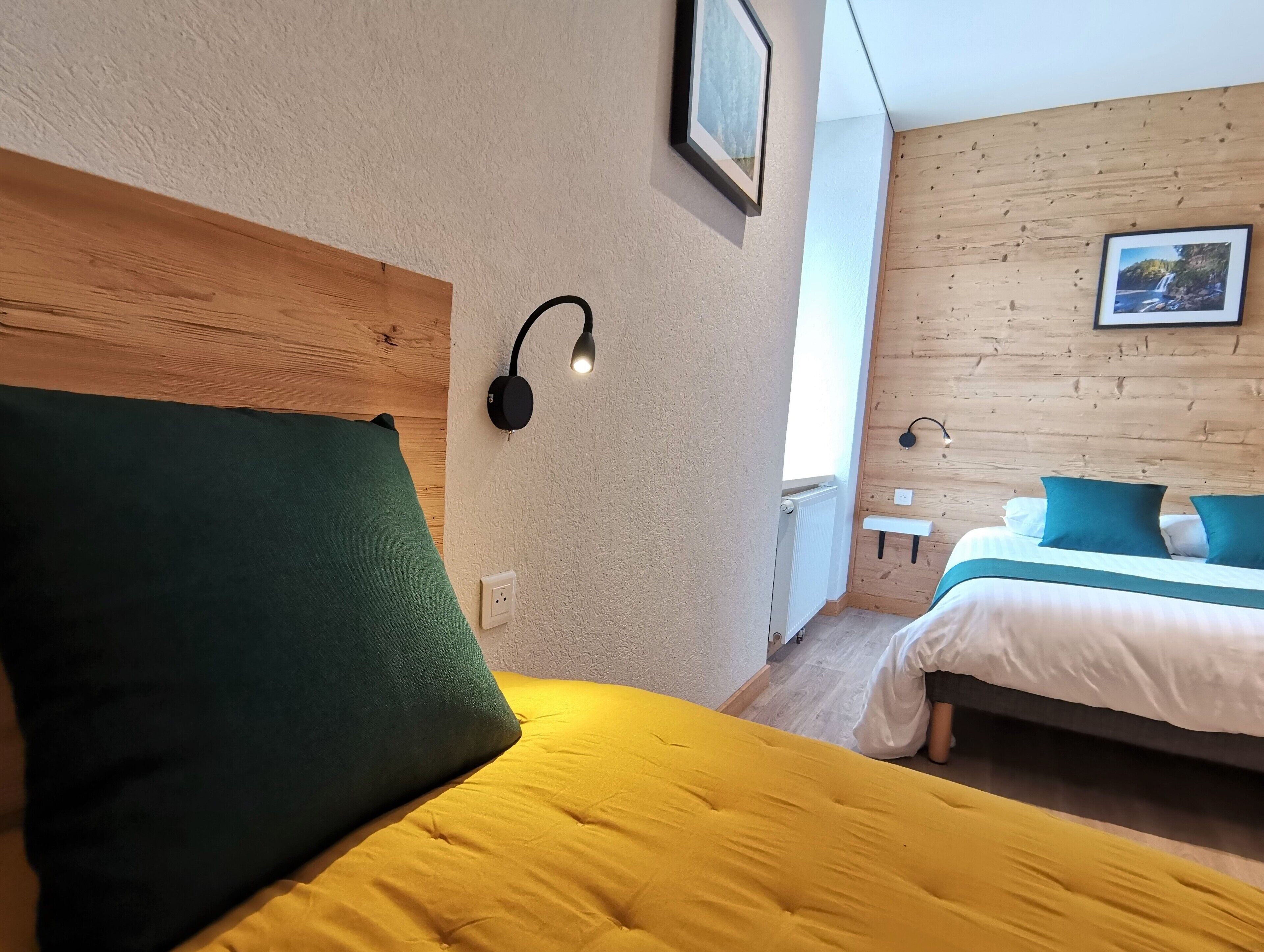 A hotel room with a wooden headboard, a green pillow, a yellow bedspread, a bedside table with a lamp, and a bed with white and teal bedding.