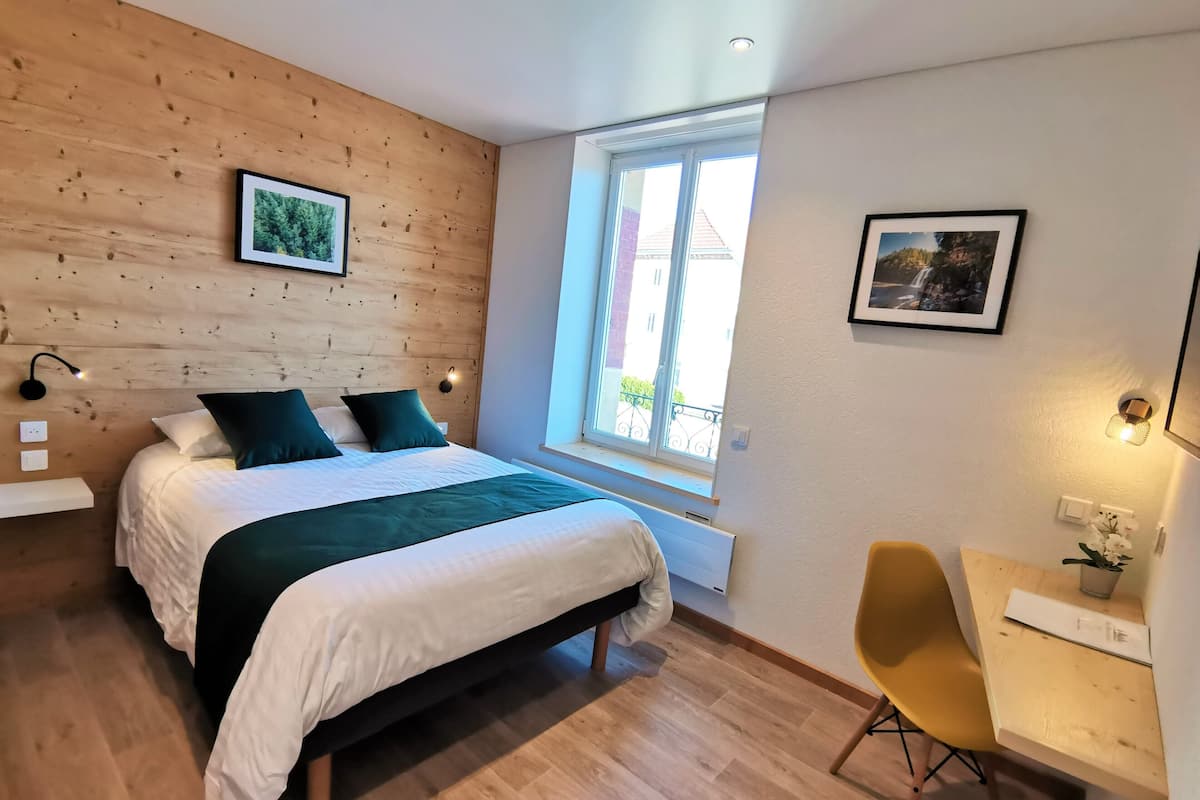 Une chambre moderne avec une tête de lit en bois, un grand lit avec une literie blanche et des coussins turquoise, un bureau avec une chaise et une fenêtre donnant sur des immeubles.