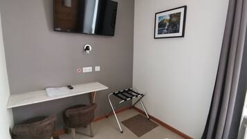 A hotel room with a flat-screen TV mounted on the wall, a small desk with a chair, a coat rack, and a bed with a blue blanket.