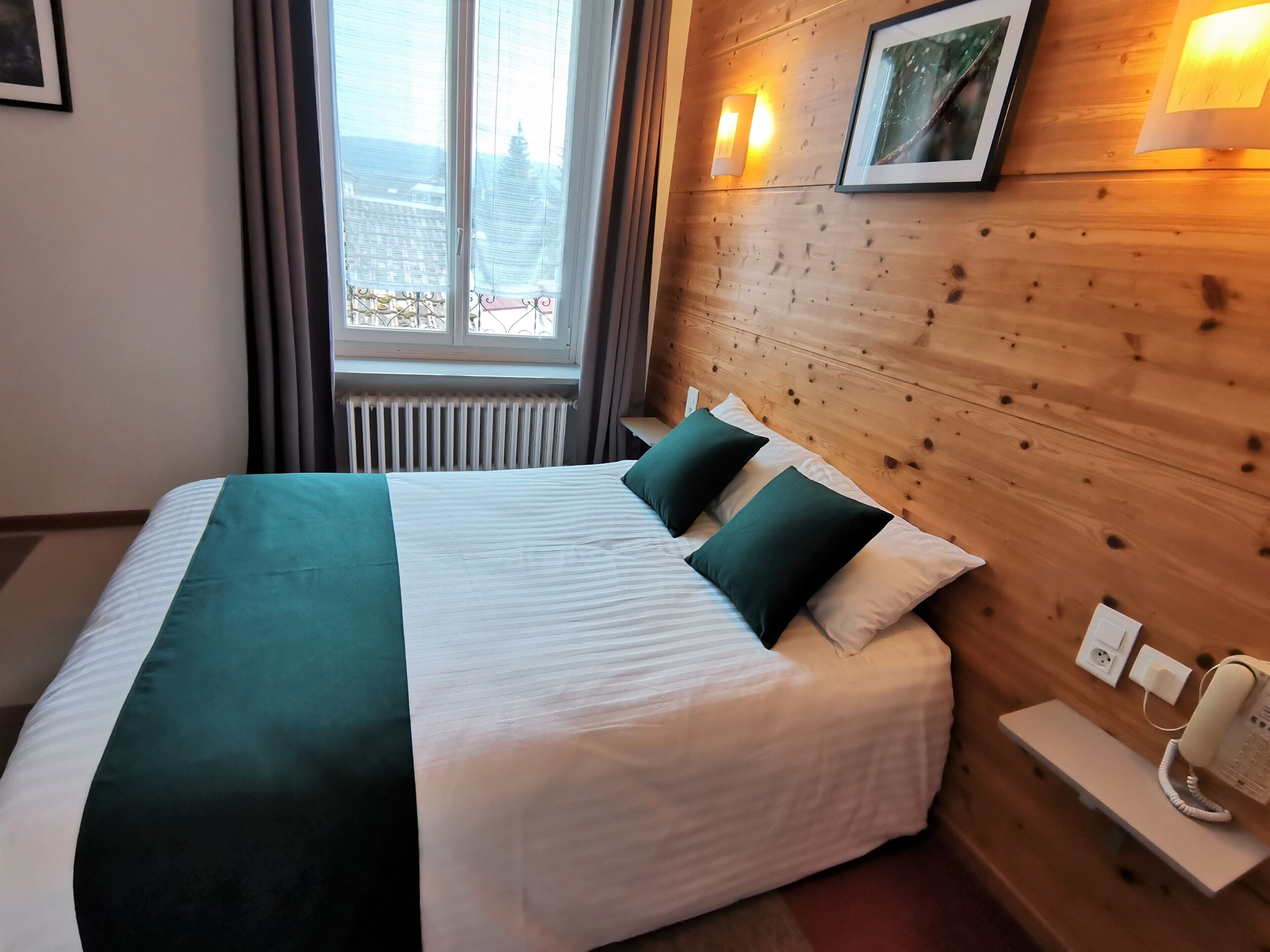 Habitación de hotel con una cama grande, un cabecero de madera, una mesita de noche con teléfono y una ventana con cortinas.