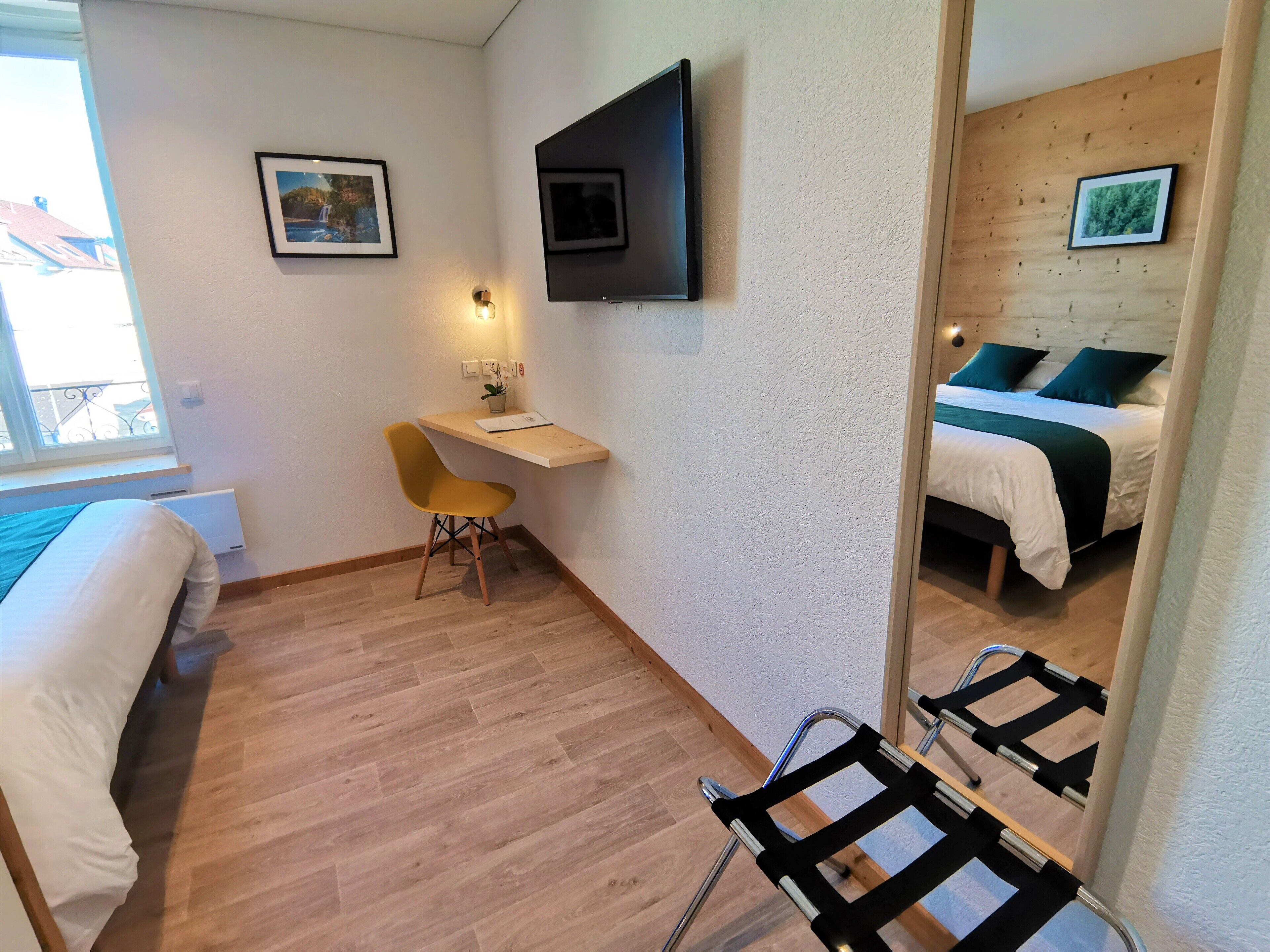 A compact hotel room with a bed, a desk with a chair, a TV mounted on the wall, and a window providing natural light.