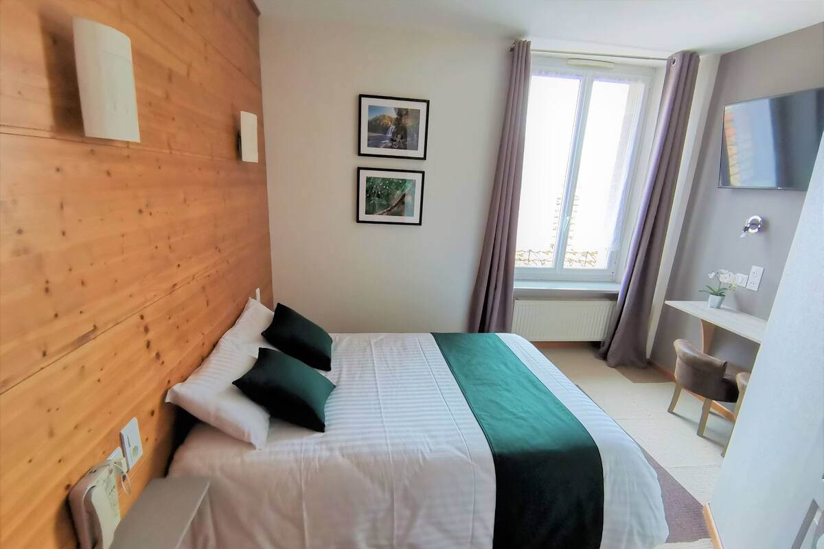 Une chambre d’hôtel avec une tête de lit en bois, un lit avec une literie blanche et verte, deux tableaux encadrés au mur, une fenêtre avec des rideaux, un bureau avec une chaise et un téléviseur à écran plat.