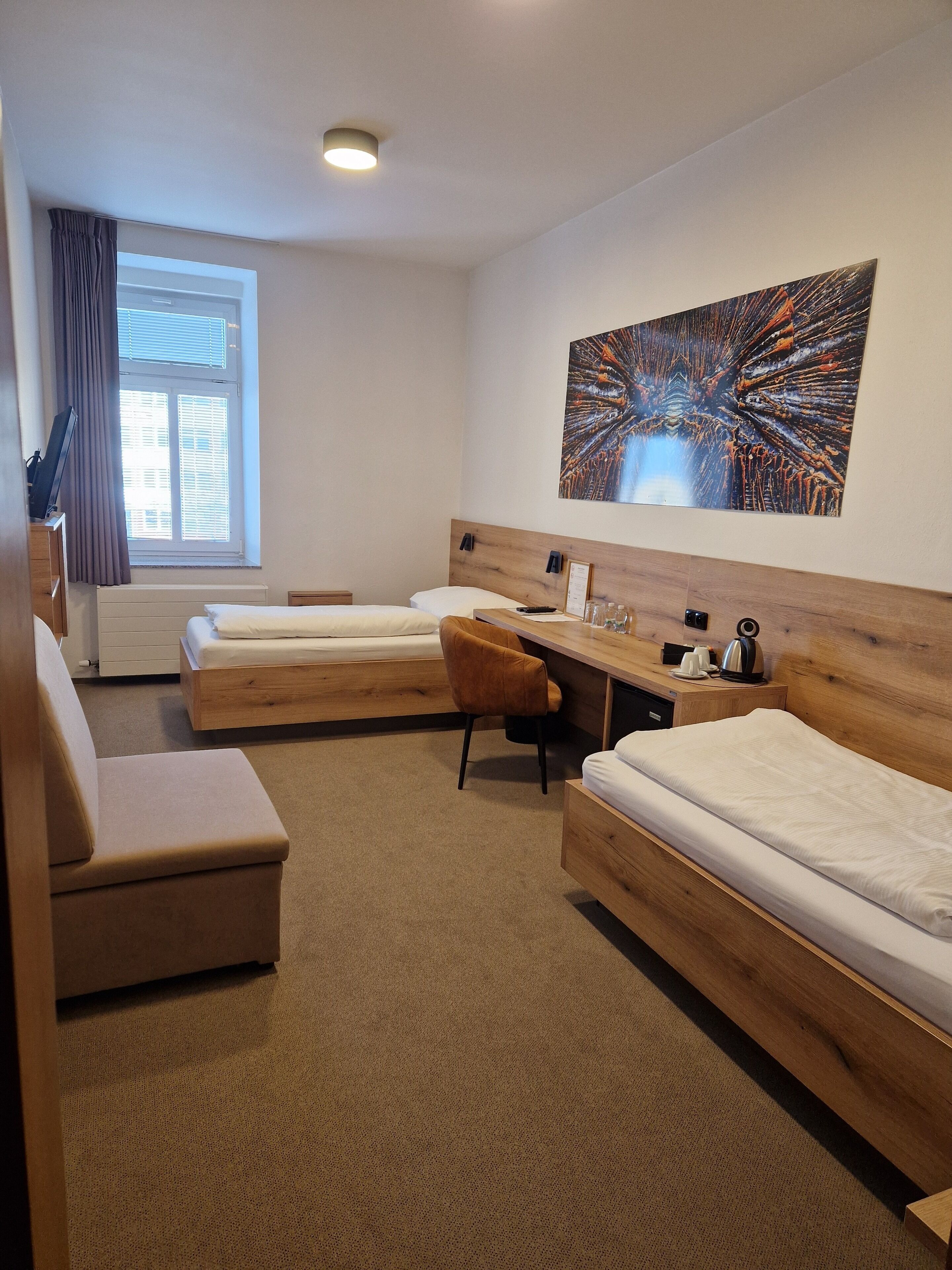 Une chambre d’hôtel avec deux lits simples, un bureau en bois, une chaise, une grande fenêtre avec des rideaux et une œuvre d’art accrochée au mur.