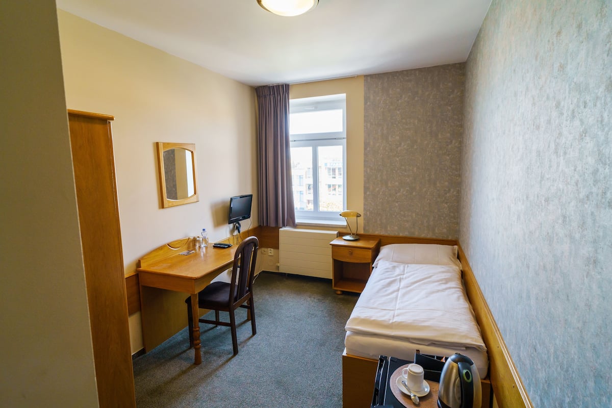 Ein Hotelzimmer mit einem Einzelbett, einem Schreibtisch mit Stuhl, einem Fernseher und einem Fenster mit Vorhängen.