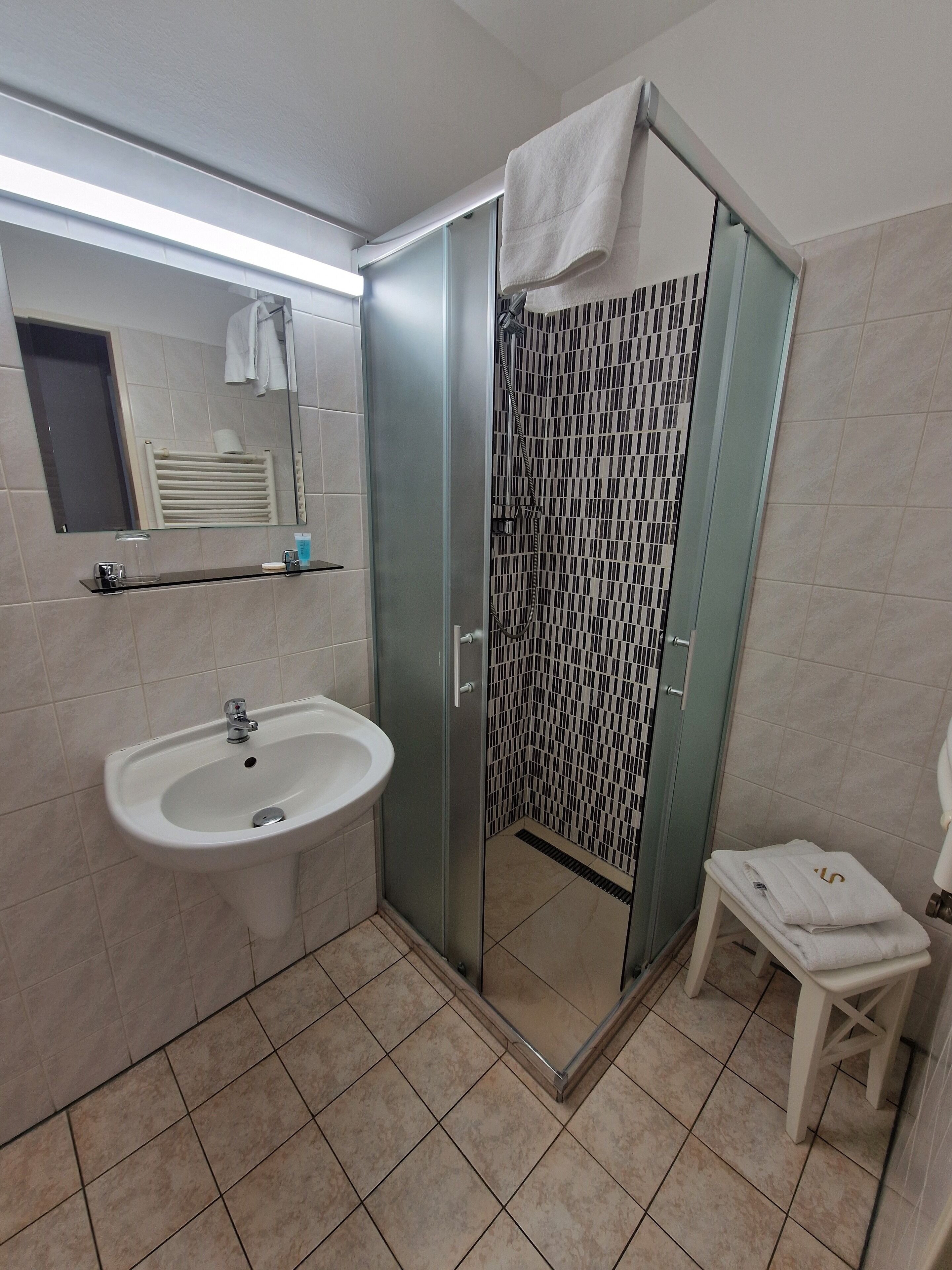 Un baño con ducha de vidrio, un lavamanos y un taburete pequeño.