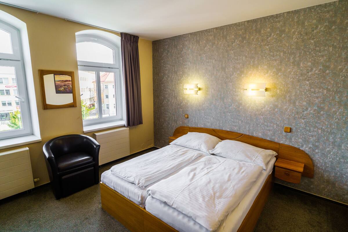 Ein Hotelzimmer mit Doppelbett, einem Sessel, einem Fenster mit Vorhängen und zwei Wandleuchten.