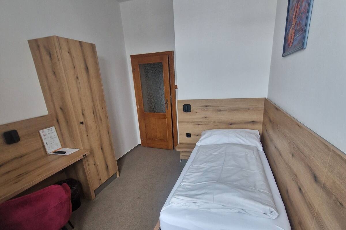 Ein kleiner, ordentlicher Raum mit einem Einzelbett, einem Holztisch, einem Stuhl und einem Kleiderschrank.