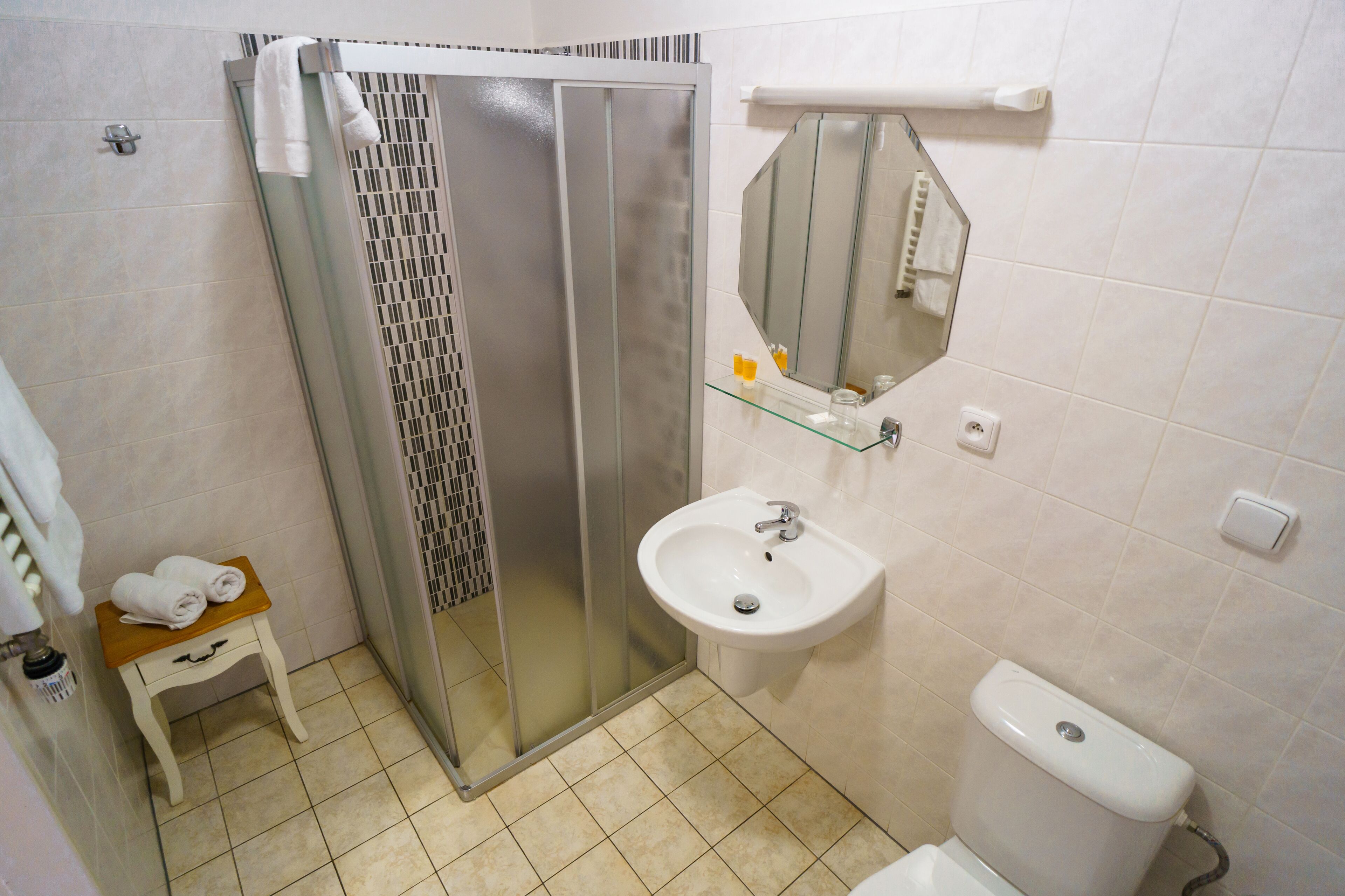 Un baño con cabina de ducha de vidrio, un lavabo, un inodoro y un taburete de madera pequeño con toallas.