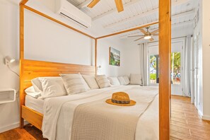 A bedroom with a wooden headboard, white bedding, a woven hat on the bed, and a view of the outdoors through a door.