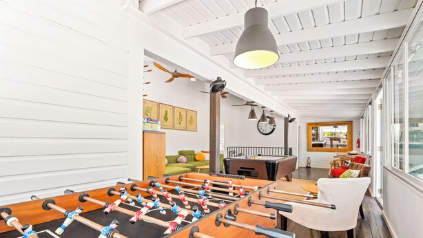 A room with a foosball table, a sofa, a dining table, and a view through a window.