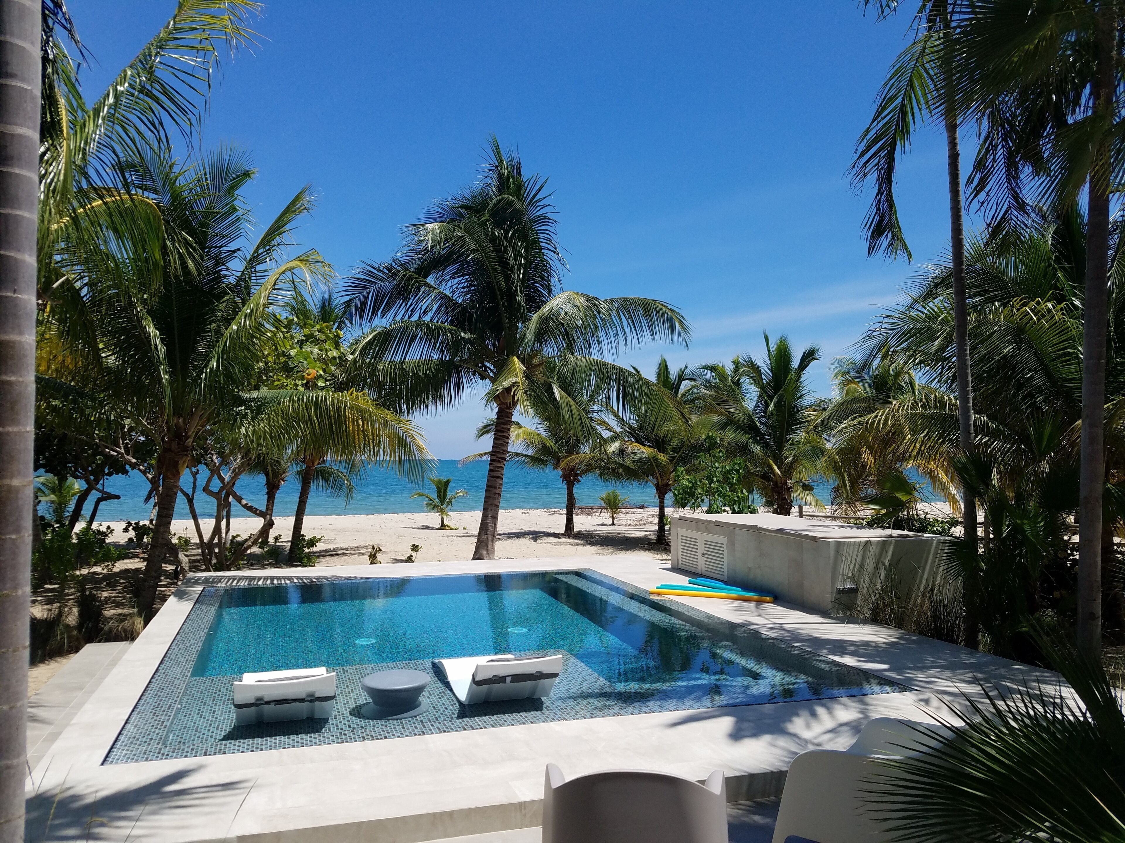 A pool area with lounge chairs, a clear blue sky, and a beach in the background.