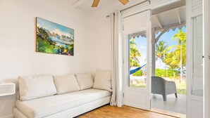Une pièce avec un canapé blanc, un tableau représentant un paysage tropical et une vue sur des palmiers à travers une porte vitrée.